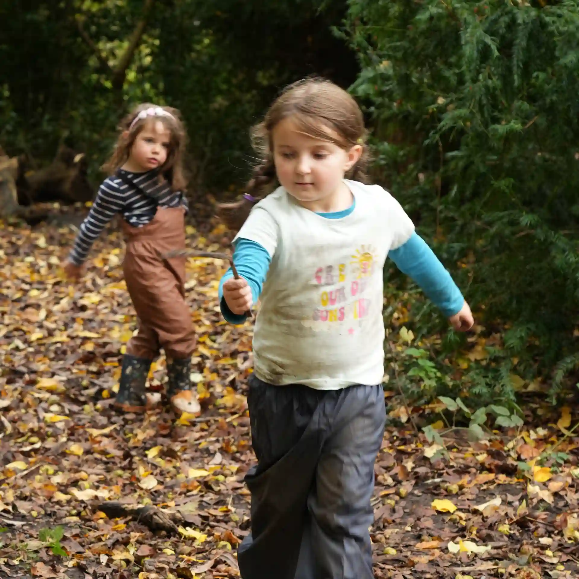 Two children running through the forest playing with sticks.